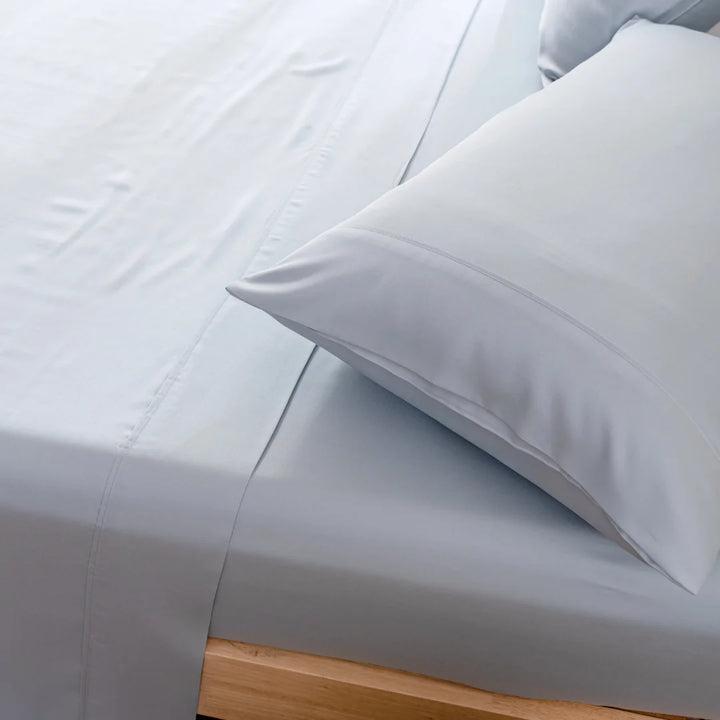 Crisp and clean bedding with neatly tucked sheets and fluffed-up pillows encased in a Linenly organic bamboo Pale Blue pillowcase set on a modern wooden bed frame, inviting a peaceful and comfortable rest.