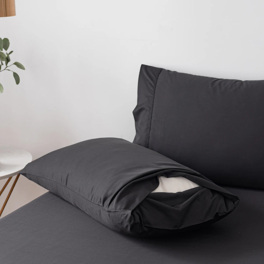A simple, elegant bedroom setup with Linenly black bamboo pillowcases on pillows, resting against a matching sheet, with a white wall backdrop and a plant on a side table adding a touch of greenery.