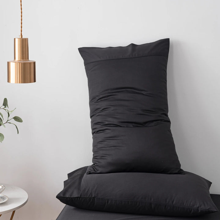 A Linenly Bamboo Pillowcase Set in Black with luxury comfort resting against a white wall, with a brass-colored pendant light hanging in the upper left corner and a hint of greenery from an indoor plant in the background.