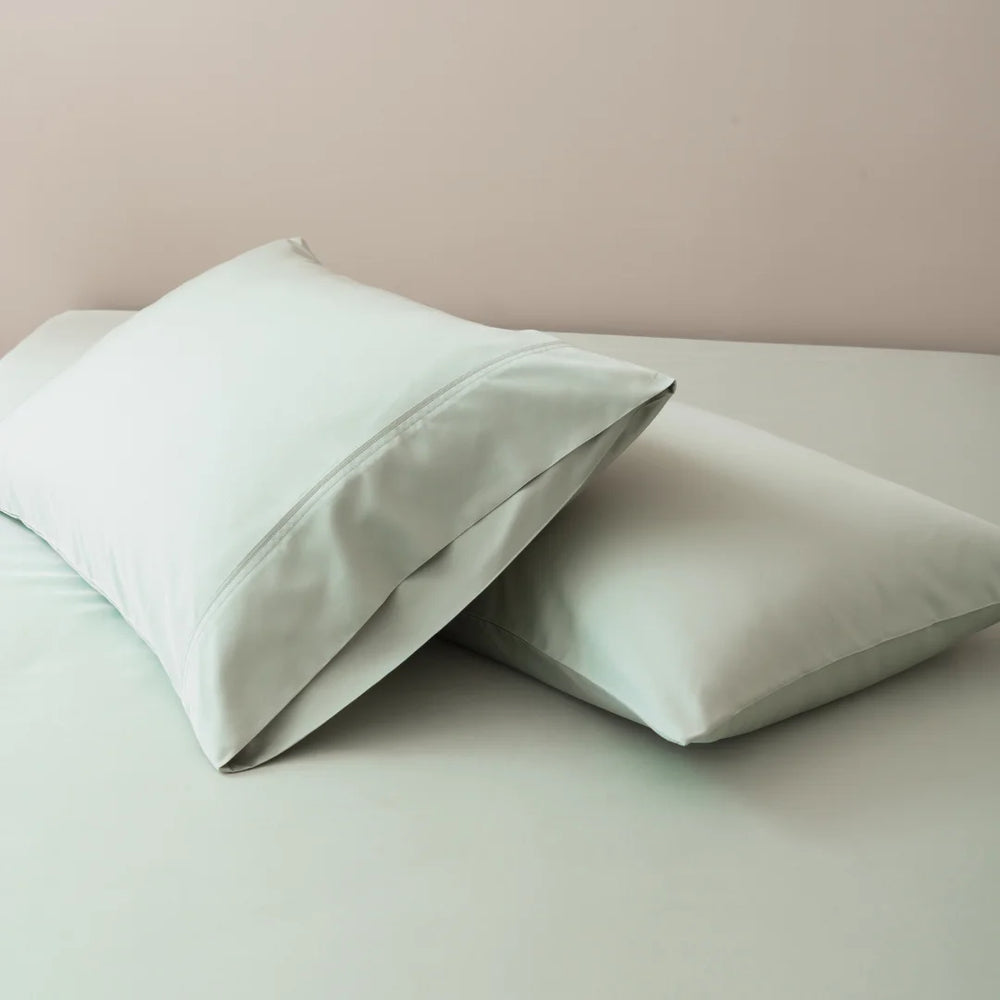 Two plush pillows with Linenly organic bamboo pillowcase set in Sage rest on a neatly made bed with smooth pastel bedding.