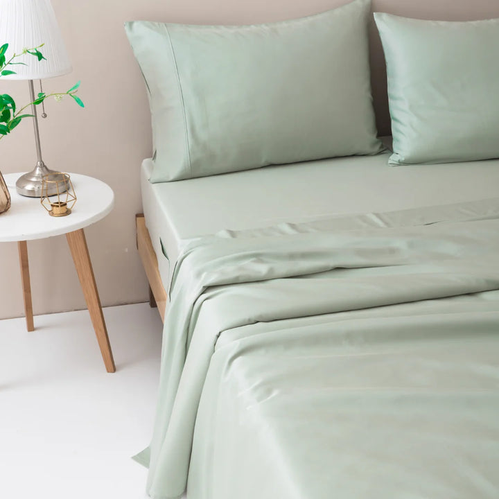 A neatly made bed with soft green sheets in a tranquil bedroom setting, accompanied by a small wooden bedside table with a lamp and a Linenly organic bamboo hypoallergenic pillowcase in sage.