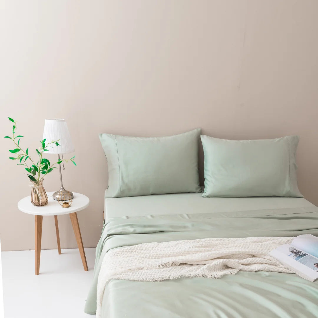 A neatly made bed with soft green linens and a Linenly organic bamboo pillowcase set in sage in a minimalist bedroom, accompanied by a small wooden bedside table holding a lamp and a glass vase with fresh greenery.