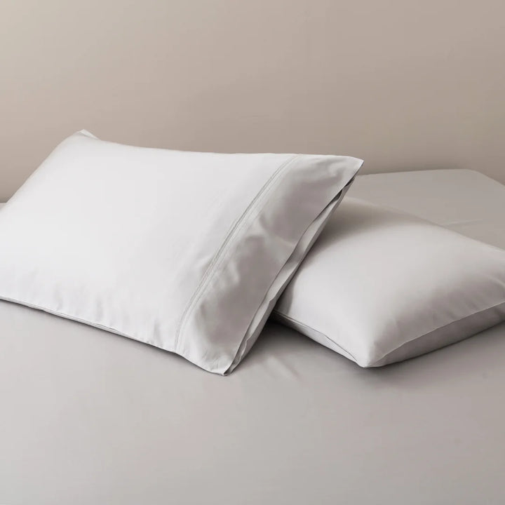 Two white pillows with Linenly Organic Bamboo Pillowcase Set - Silver on a beige surface.