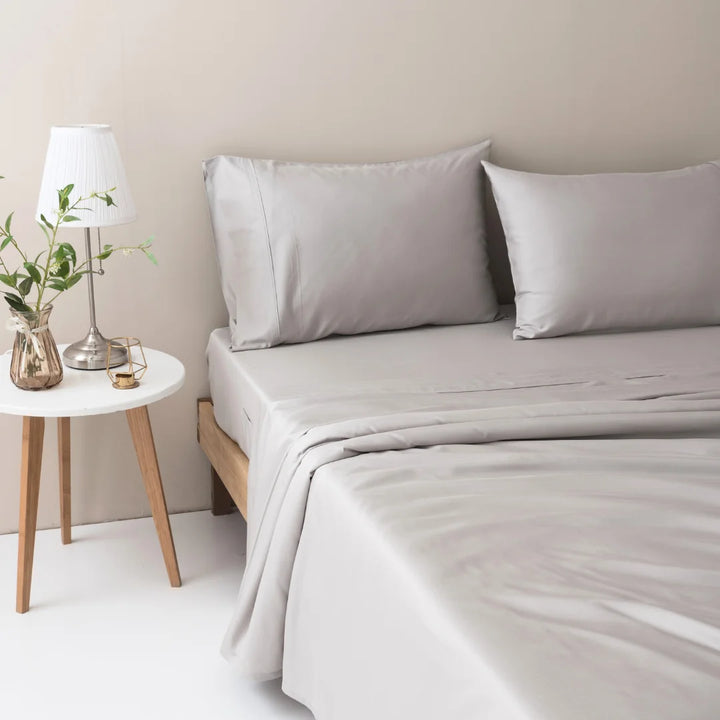 A tranquil and neatly made bed with soft gray linens featuring Linenly organic Bamboo Pillowcase Set in Silver, complemented by a wooden bedside table featuring a white lamp and a clear vase with green foliage.