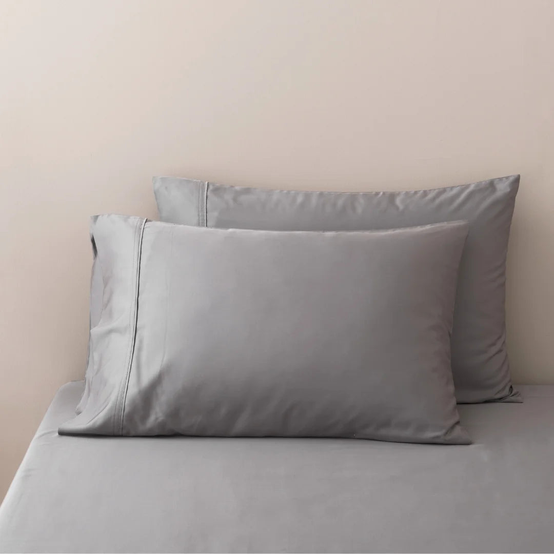 A neatly made bed with a set of Linenly Stone Grey Bamboo Pillowcases on a matching gray bedsheet against a light beige wall.