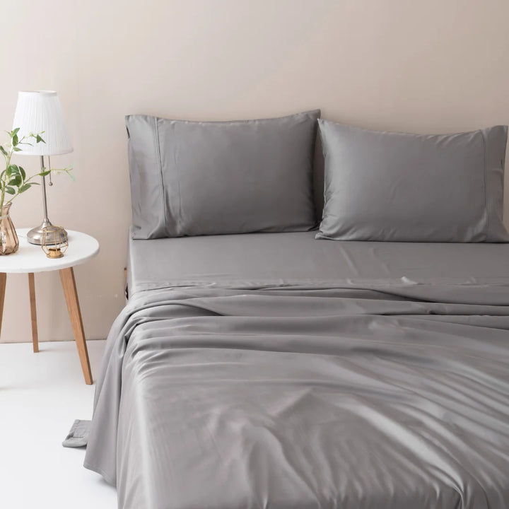 A neatly made bed with grey sheets and Linenly bamboo pillowcase set in Stone Grey in a tranquil bedroom setting, accompanied by a wooden bedside table with a white lamp and a vase with fresh plants.
