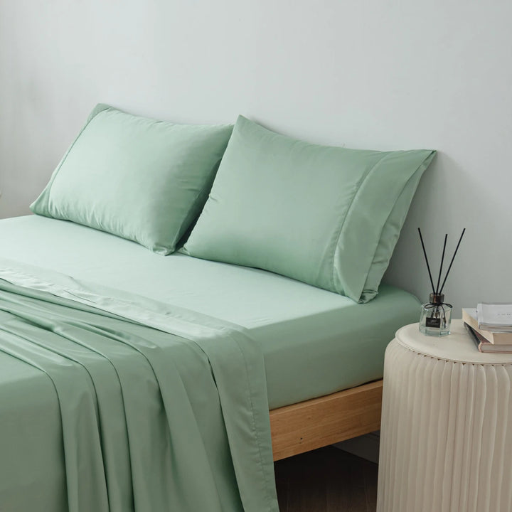 A neatly made bed with smooth, sage green bedding, including a fitted sheet, a flat sheet, and two Linenly Bamboo Pillowcase Sets in Summer Green, in a minimalist bedroom setting with a subtle aroma diffuser on the side.