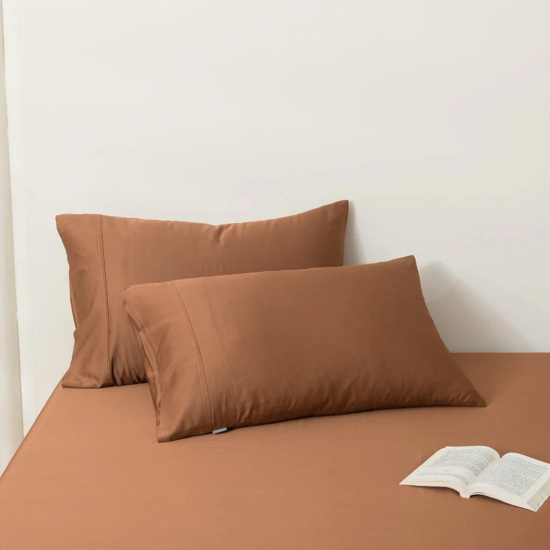 Two brown Linenly bamboo pillowcases resting against a white wall on a matching brown surface, with an open book placed in front of them, inviting a moment of relaxation and reading.