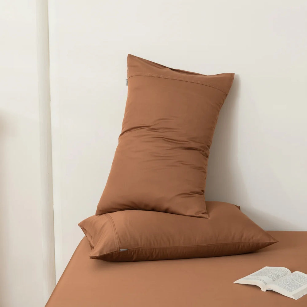 A minimalist setting with an earth-toned, Linenly Bamboo Pillowcase Set in Terracotta leaning against a white wall on a matching bench with a casually placed open book, suggesting a quiet, cozy reading nook.
