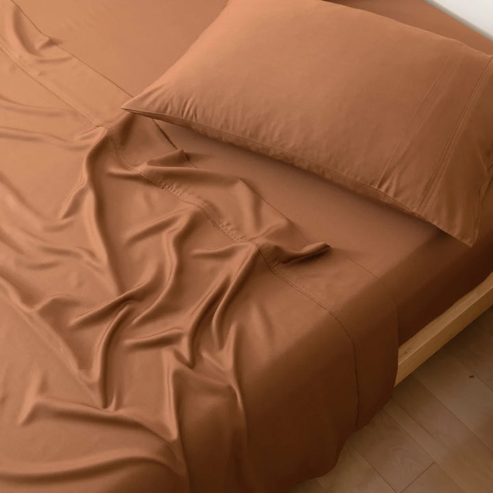 A neatly made bed with smooth terracotta-colored linens and Linenly Terracotta Bamboo Pillowcase Set in a warmly lit room.