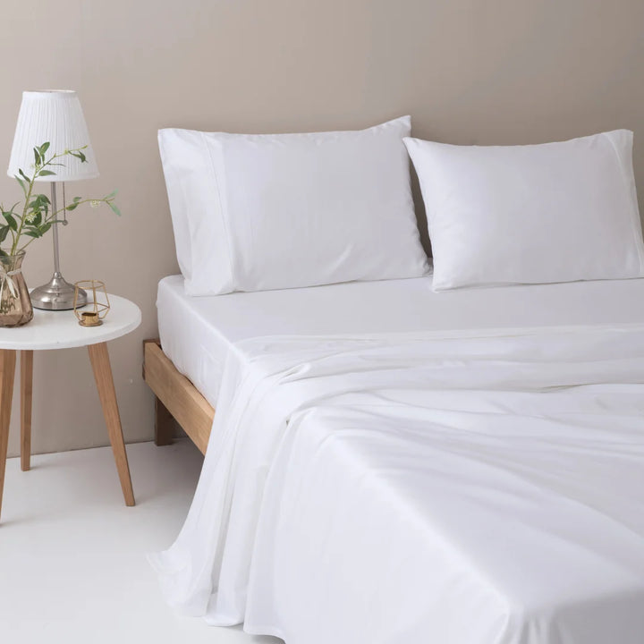 A neatly made bed with white linens and Linenly organic Bamboo Pillowcase Sets in white, accompanied by a wooden bedside table featuring a lamp and a vase with greenery to create a serene and minimalist bedroom setting.