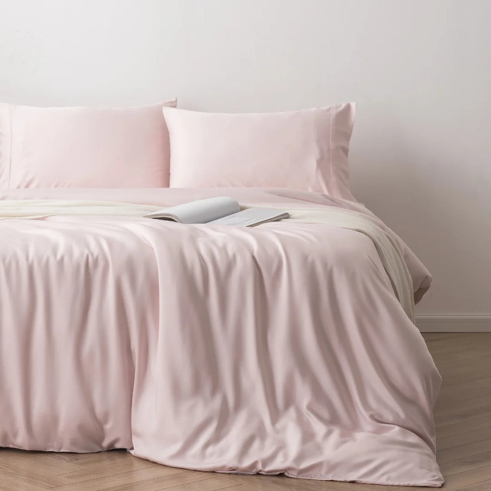 A neatly made bed with a soft pink Linenly Bamboo Quilt Cover - Blush and a pair of pillows against a light grey wall provides a luxurious sleep experience.