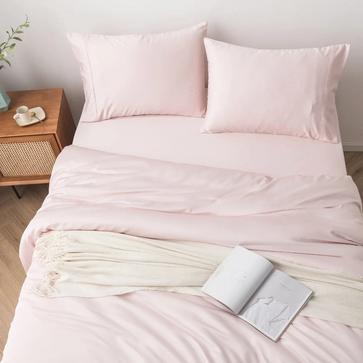 A neatly made bed with soft pink Linenly Blush Bamboo Quilt Cover bedding and a cozy cream throw blanket at the end, accompanied by an open book, inviting a peaceful reading session.