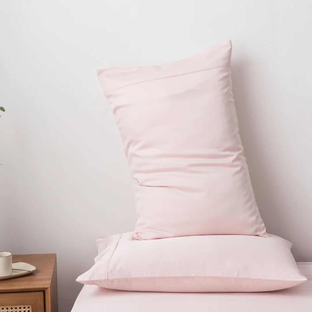 A soft pink pillow standing upright on a neatly made bed with a minimalist wooden bedside table and a simple white wall in the background, covered by a Linenly Blush Bamboo Quilt Cover for a more luxurious sleep.