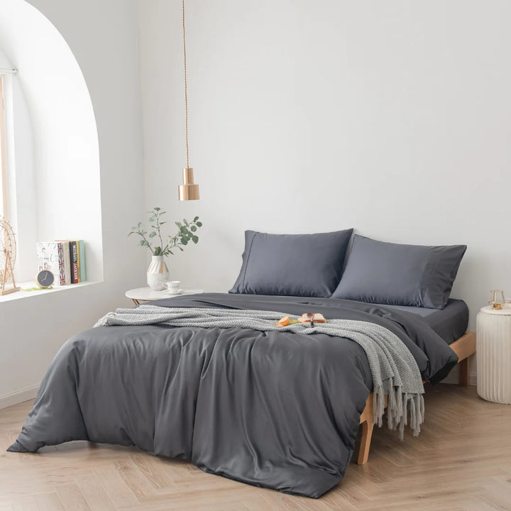 A neatly made bed with a Linenly eco-friendly Charcoal Bamboo Quilt Cover in a bright, minimalist bedroom, complemented by a small bedside table with books and a plant, and a cozy grey throw blanket rested at the end.
