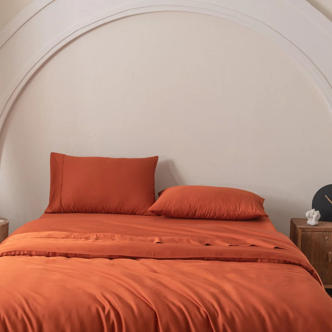 A Bamboo Quilt Cover in Copper Sunset by Linenly, crafted from premium bamboo, with two pillows adorns a bed against a white wall featuring a large, arched design. A wooden nightstand with a decorative object is visible beside it, giving the room a minimalist and cozy aesthetic.