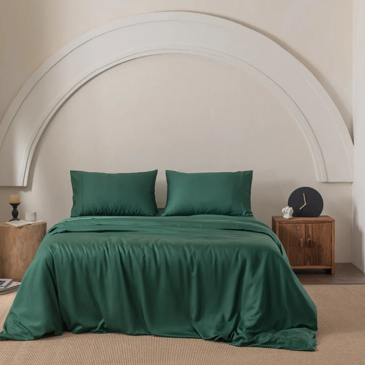 In a bedroom decorated with a Linenly Bamboo Quilt Cover Set - Evergreen, a bed adorned with green pillows sits against an arch-designed white wall. A wooden nightstand holds a lamp, candle, and clock. The premium bamboo materials provide silky comfort, while a textured rug completes the room.