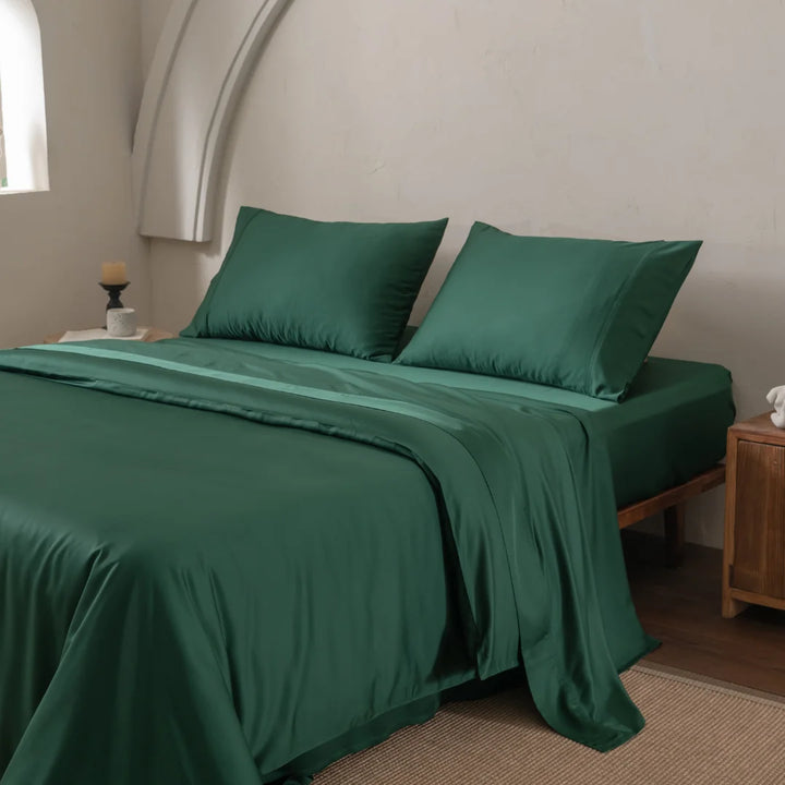 A neatly made bed with the Bamboo Quilt Cover Set in Evergreen by Linenly features deep green, silky-textured sheets and pillows against a light wall. A small wooden nightstand and window add to the cozy, elegant bedroom atmosphere, enhanced by premium bamboo materials.