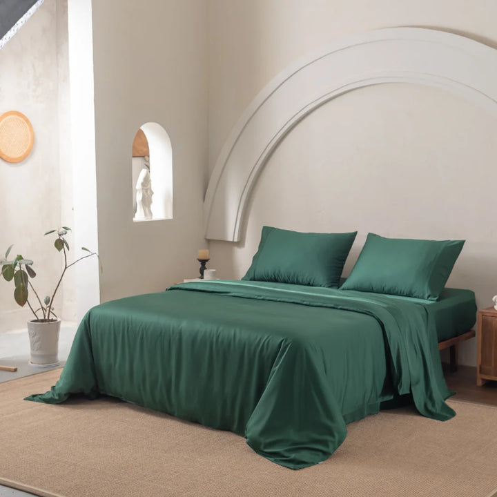 The Bamboo Quilt Cover Set - Evergreen by Linenly transforms a bedroom into a restful retreat with its green bedspread on a large bed. The room features an arched beige wall, small potted plant, wooden side table, and wicker decor, all enhanced by silky textures and bright natural light.