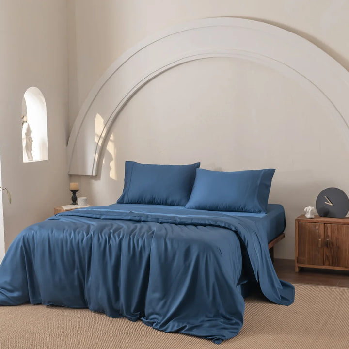 A cozy bedroom showcases a bed adorned with Linenly's Bamboo Quilt Cover in Midnight, offering a silky feel beneath a decorative white arch. To the right, a wooden nightstand holds a lamp and clock, while natural light from a small window illuminates the floor rug.