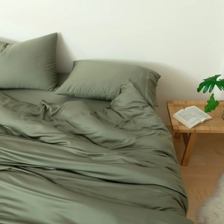 A comfortable and neatly made bed with a smooth green Bamboo Quilt Cover - Moss crafted from environmentally friendly organic bamboo viscose by Linenly, a wooden bedside table featuring an open book, and a hint of greenery to create a calming bedroom