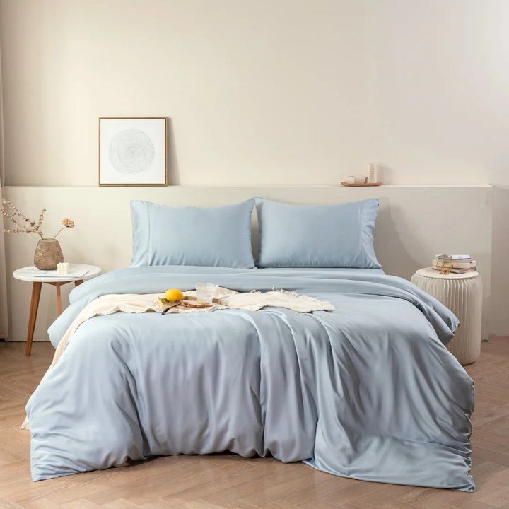 A neatly made bed with a Linenly pale blue bamboo quilt cover in a serene bedroom setting, featuring a simple white bedside table with decorative items and a framed artwork on the wall.