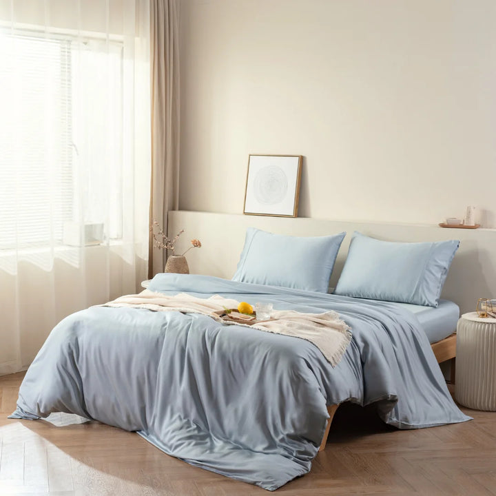 A serene bedroom with a neatly made bed featuring a Linenly pale blue bamboo quilt cover with anti-sweat properties, a cozy beige throw, a wooden bedside table, and a calming, natural light ambiance.