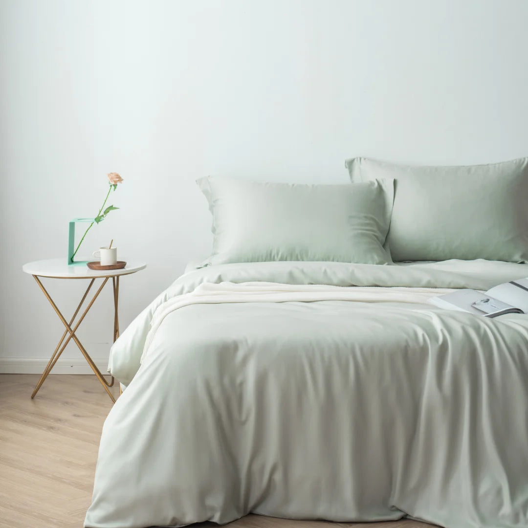 A neatly made bed with soft green linens, specifically a Linenly sage bamboo quilt cover in a minimalist bedroom, accompanied by a small side table with a single rose in a vase and a book, evoking.