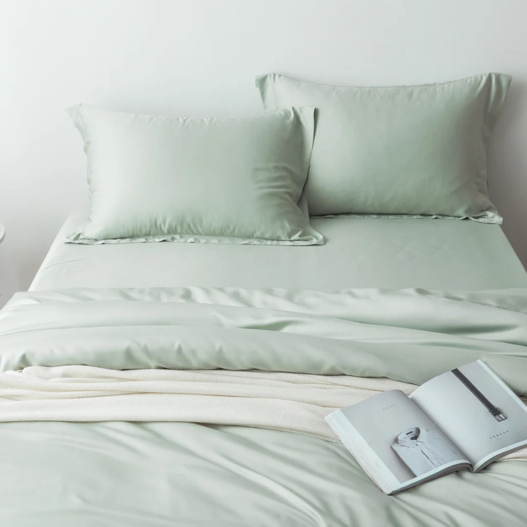 A neatly made bed with a Linenly sage bamboo quilt cover and a cozy cream blanket, accompanied by an open magazine resting on the bed.