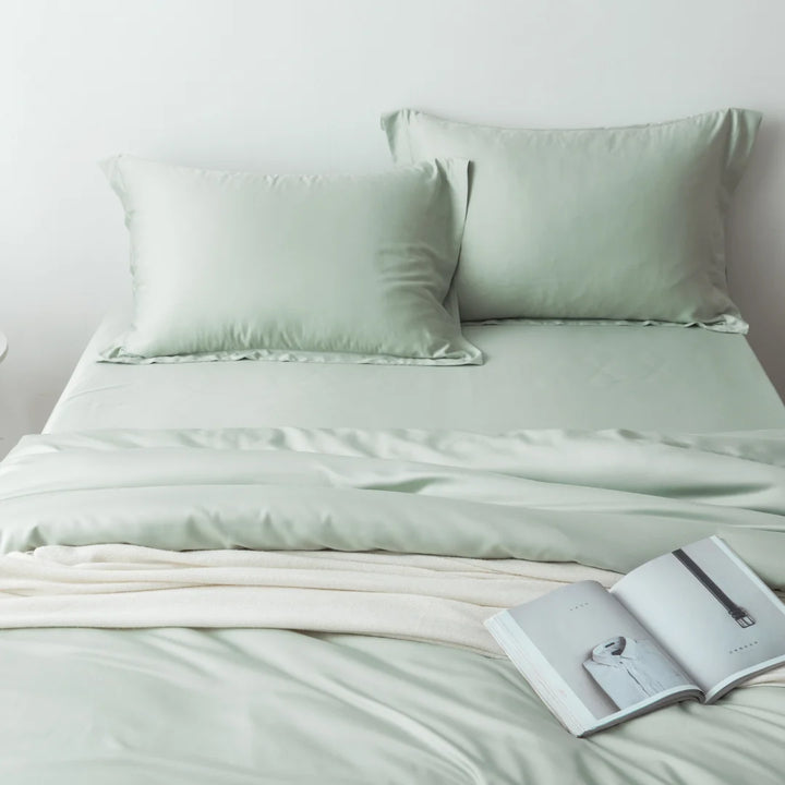 A neatly made bed with a Linenly sage bamboo quilt cover and a cozy cream blanket, accompanied by an open magazine resting on the bed.