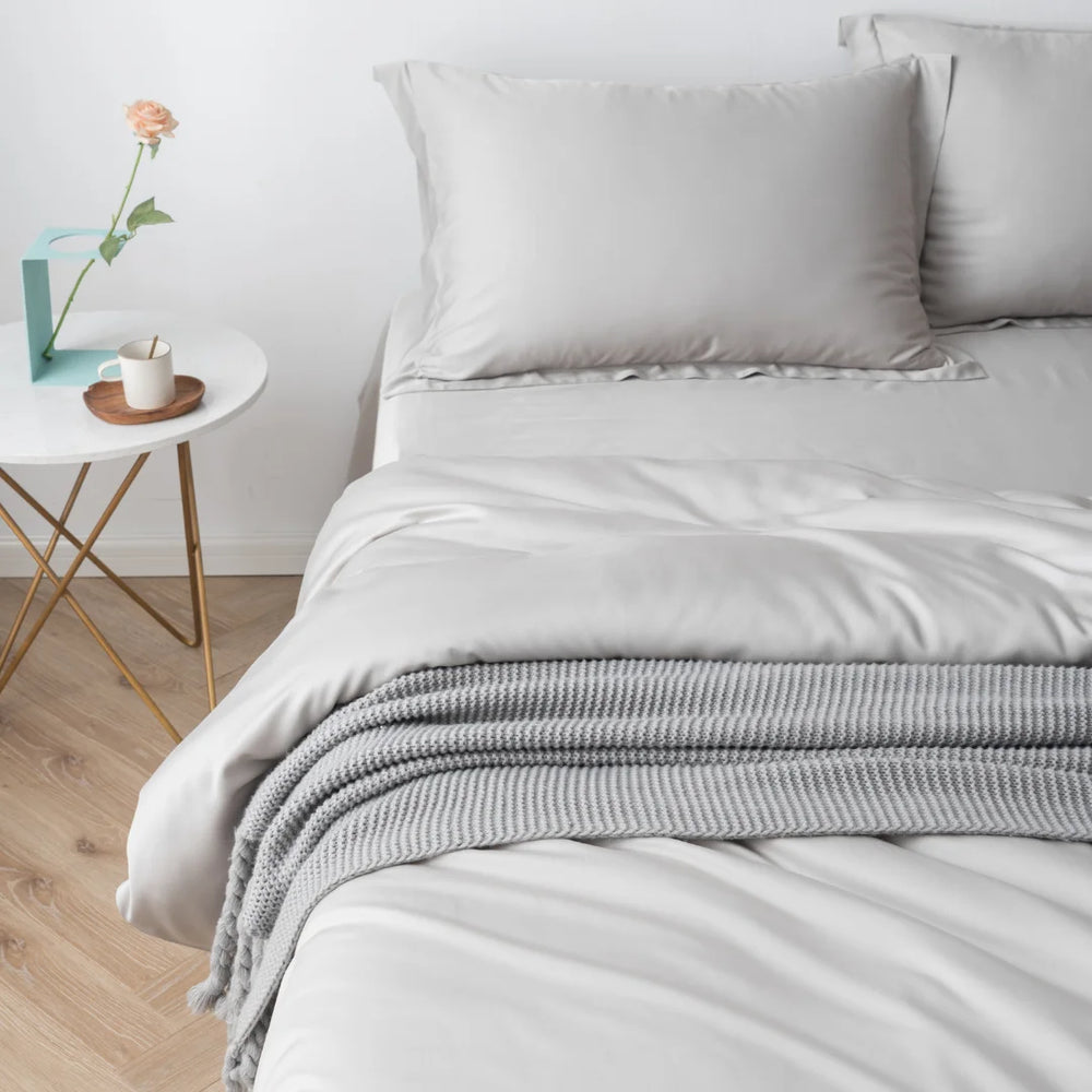 A neatly made bed with Linenly's Bamboo Quilt Cover in Silver and a gray knitted throw at the foot, beside a modern nightstand with a single rose in a vase and a cup on a saucer.