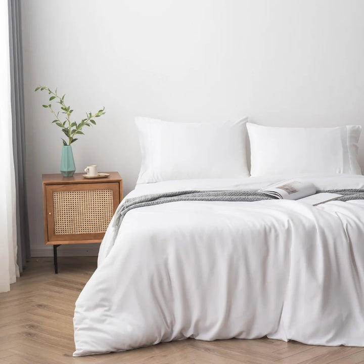 A cozy and minimalist bedroom with a neatly made bed featuring a Linenly white bamboo quilt cover with sateen weave, a wooden bedside table with a blue vase and greenery, a small dish, and a