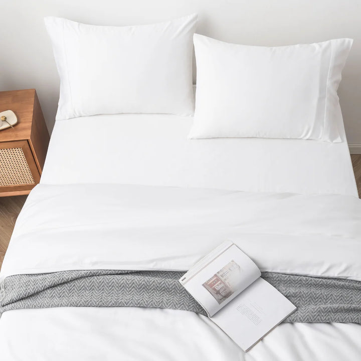A neatly made bed with a Linenly crisp white bamboo quilt cover and an open book lying on the grey textured blanket at the foot of the bed, signifying a quiet, peaceful moment in a bright, clean bedroom.