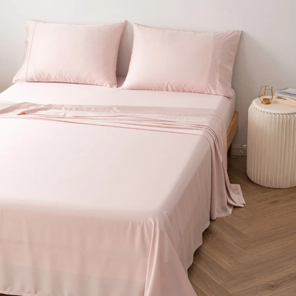 A neatly made bed with Linenly's ultra-soft Bamboo Sheet Set in Blush and pillowcases in a tranquil bedroom setting, accompanied by a simple bedside table and a hardwood floor.