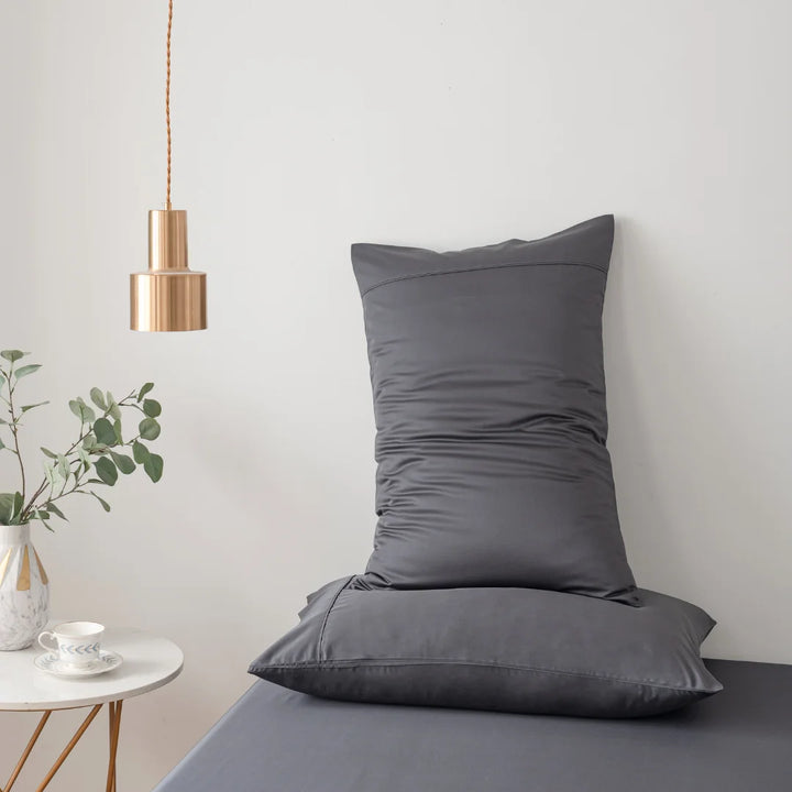 A minimalist and serene corner with a sleek gray cushioned chair, accompanied by a small vase with greenery, a chic hanging copper lamp, and a simple white cup on a saucer atop a luxurious Linenly Bamboo Sheet Set in Charcoal.