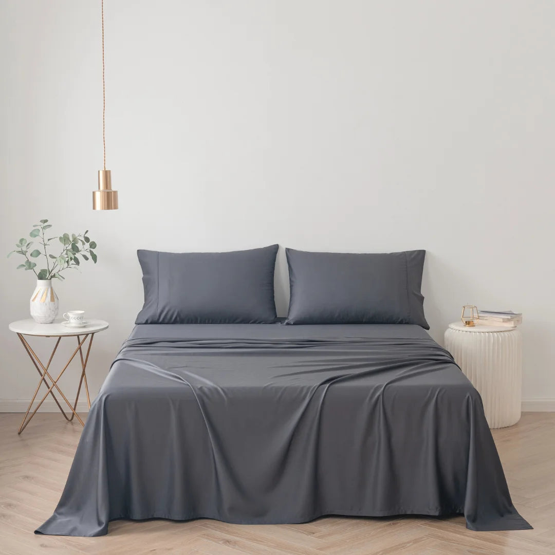 A modern minimalist bedroom with a neatly made bed featuring a luxurious comfort Linenly Charcoal Bamboo Sheet Set, complemented by a simple white side table with an eco-friendly living plant, and an elegant copper pendant light.