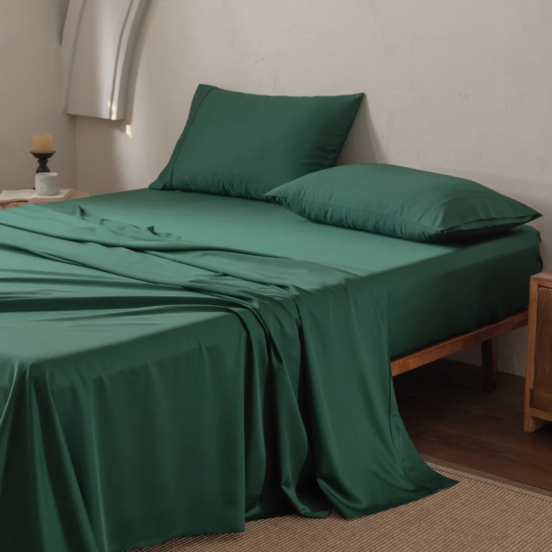 A neatly made bed features the ultra-soft, silky-smooth Bamboo Sheet Set - Evergreen by Linenly, paired with matching dark green pillows in a simple room. A wooden nightstand holds a small candle and a book. The floor is covered with a beige rug as soft natural light illuminates the scene.