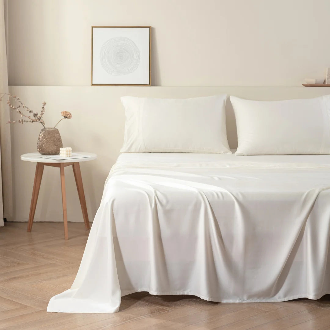 A minimalist bedroom with a neatly made bed draped in Linenly's Ivory Bamboo Sheet Set, beside a small wooden side table with a vase and decorative objects, under a framed abstract wall art.