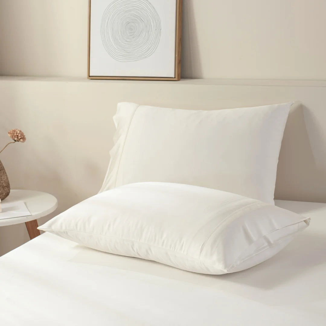 A pair of white pillows on a neatly made bed, dressed in luxury bedding including a Linenly ivory bamboo sheet set, with a minimalist side table and framed artwork in the background, creating a serene and cozy bedroom