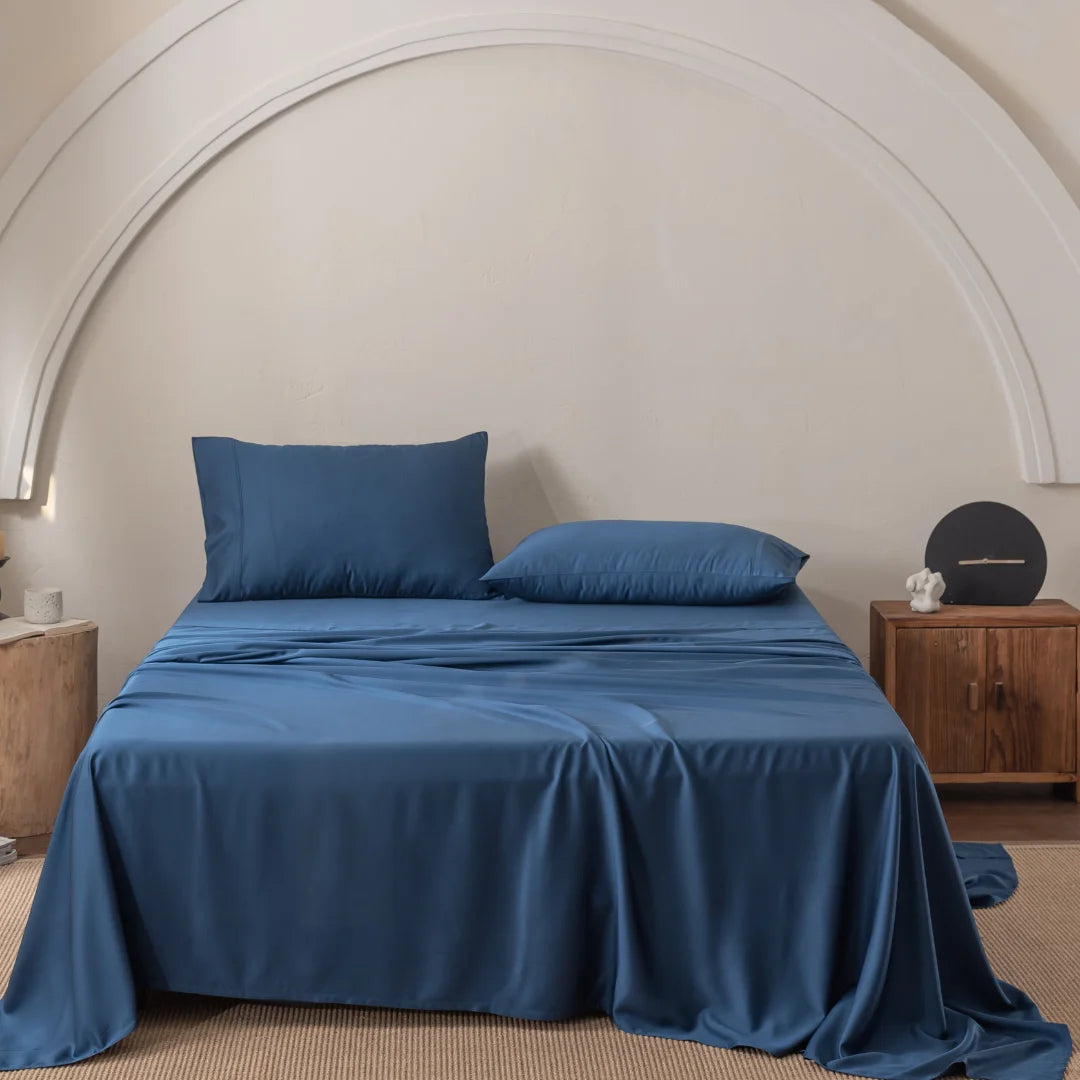 The Bamboo Sheet Set - Midnight by Linenly, with ultra-soft blue sheets and pillows, centers the minimalist room. A wooden nightstand on the right holds decorative items, while a tree stump side table sits on the left. An elegant arch design adorns the wall above in this serene setting.