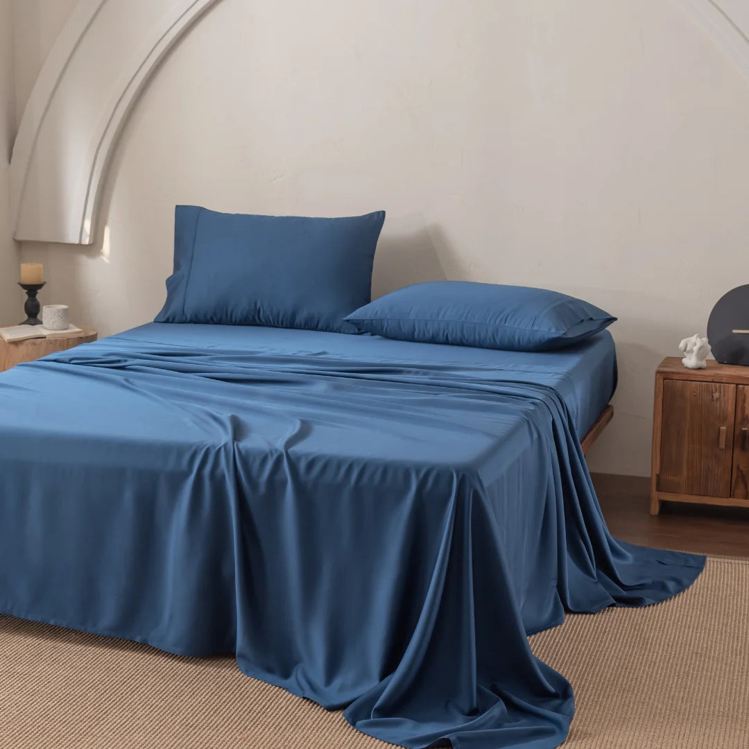 A neatly made bed with Linenly's Bamboo Sheet Set in Midnight, featuring ultra-soft dark blue bedding, two pillows, and an elegantly draped oversized blanket. The room also includes a wooden nightstand, a candle, and a neutral-colored wall with an arch design.