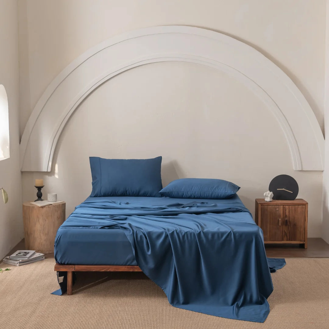 A minimalist bedroom showcases an unmade bed adorned with Linenly's ultra-soft Bamboo Sheet Set in Midnight. Two pillows rest comfortably, while a wooden nightstand holds a candle and small item. A curved white wall decor frames the bed, and beige carpet on the floor enhances the serene vibe.