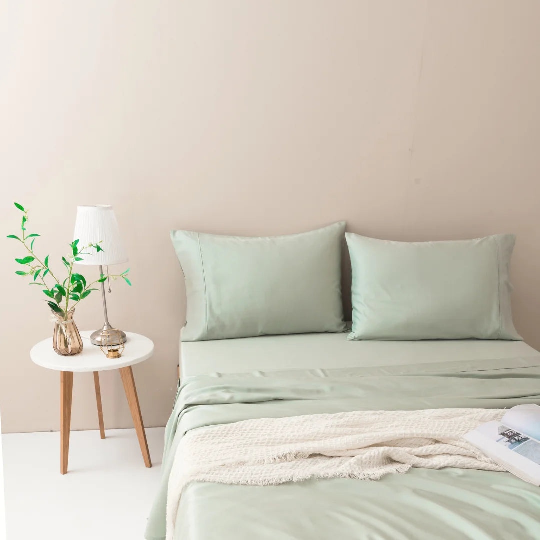 A cozy bedroom corner with a neatly made bed in pastel green, complemented by a luxuriously soft Linenly Sage Bamboo Sheet Set, created from sustainable bamboo fibers, and a small wooden side table hosting
