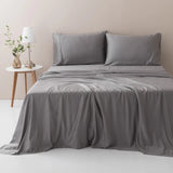 A neatly made bed with a Linenly Stone Grey Bamboo Sheet Set made from sustainable bamboo fibers in a minimalist bedroom, accompanied by a small wooden side table with a vase of greenery and a lamp.