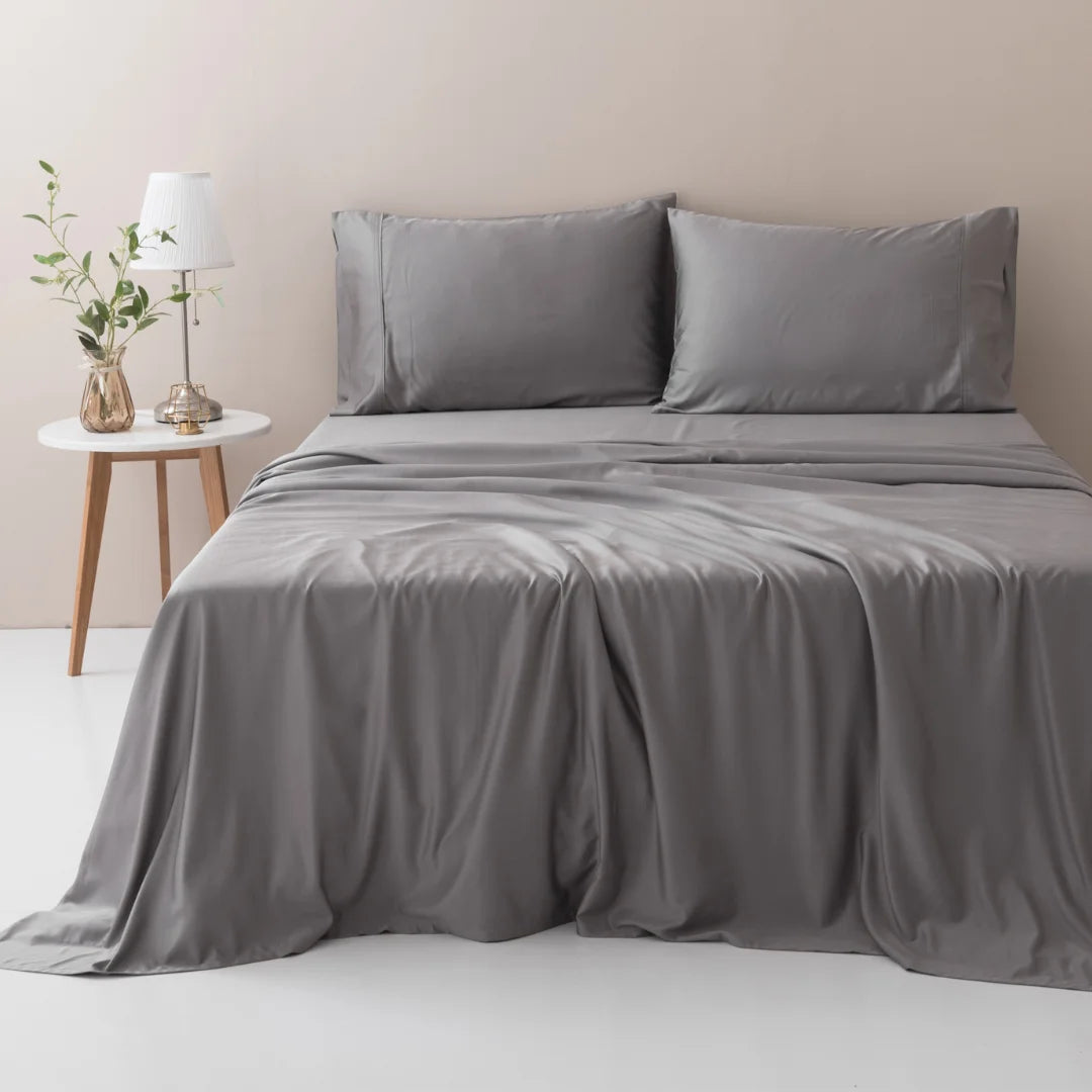 A neatly made bed with a Linenly Stone Grey Bamboo Sheet Set made from sustainable bamboo fibers in a minimalist bedroom, accompanied by a small wooden side table with a vase of greenery and a lamp.