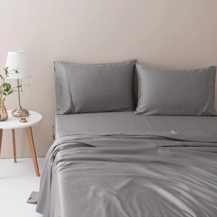 A neatly made bed with a Linenly Stone Grey bamboo sheet set in a minimalist bedroom, complemented by a white lamp and a vase with greenery on a wooden bedside table.