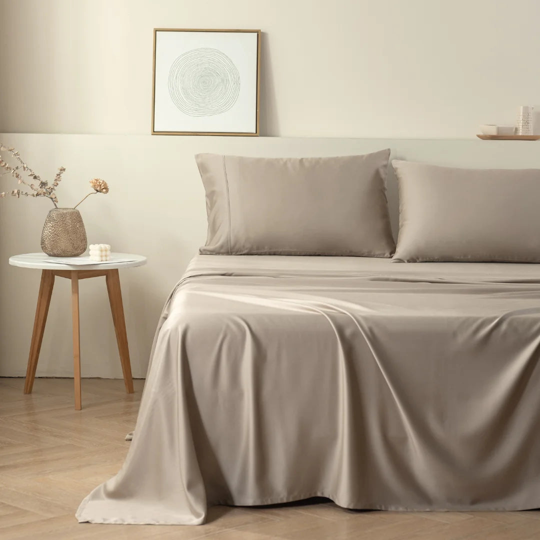 A cozy minimalist bedroom with a neatly made bed in earth tones, featuring Linenly's eco-conscious Bamboo Sheet Set in Taupe, a simplistic bedside table with a decorative object, and a framed abstract artwork on the wall, invoking