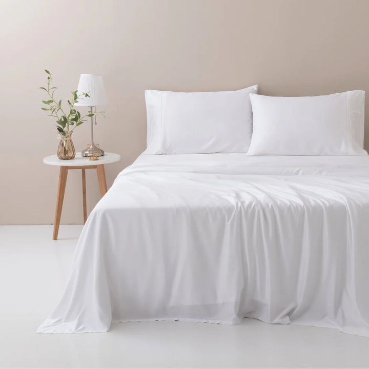 Minimalist and serene bedroom with an ultra-soft Linenly bamboo sheet set in white on a neatly made bed, complemented by a wooden bedside table with a lamp and a vase with fresh greenery.