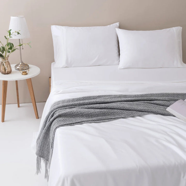 A minimalist bedroom with a neatly made bed, Linenly's Bamboo Sheet Set in White of silky-smooth texture, and a grey knitted throw blanket at the foot of the bed. A wooden bedside table with a lamp and plant.