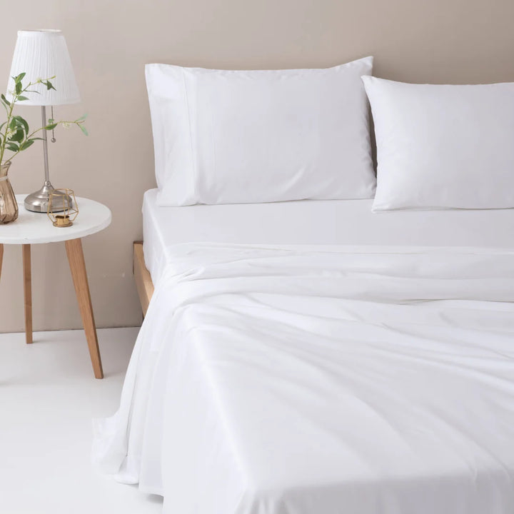 A serene and tidy bedroom setup with a neatly made bed featuring an ultra-soft Linenly bamboo sheet set in white, flanked by a wooden bedside table with a classic lamp and a vase with greenery.